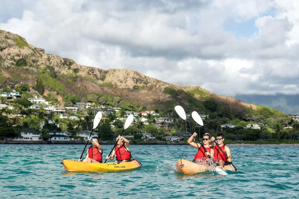 Zwei Doppelkajaks mit Paddlern in roten Schwimmwesten vor einem grünen Hang mit Häusern