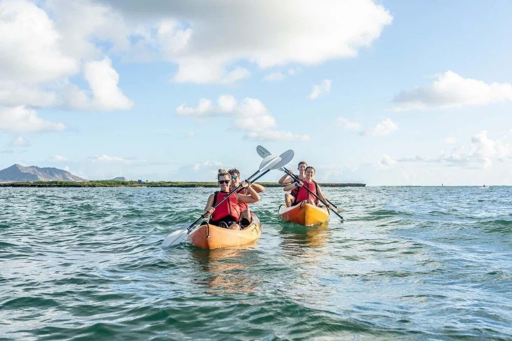 Dos kayaks dobles con palistas con chalecos salvavidas rojos avanzando por mar abierto hacia una isla baja
